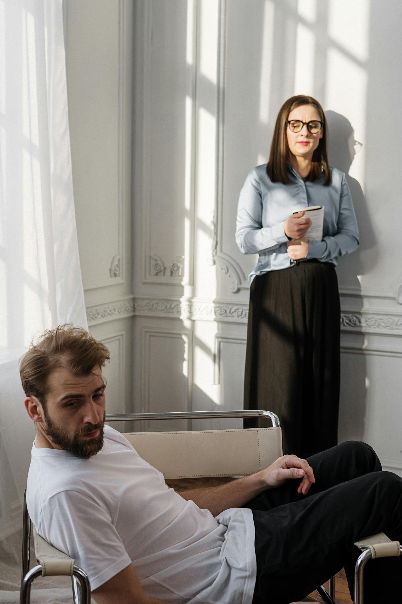 A bearded man in a white t-shirt sits in a chair while a woman holds a notepad, suggesting a counseling session.