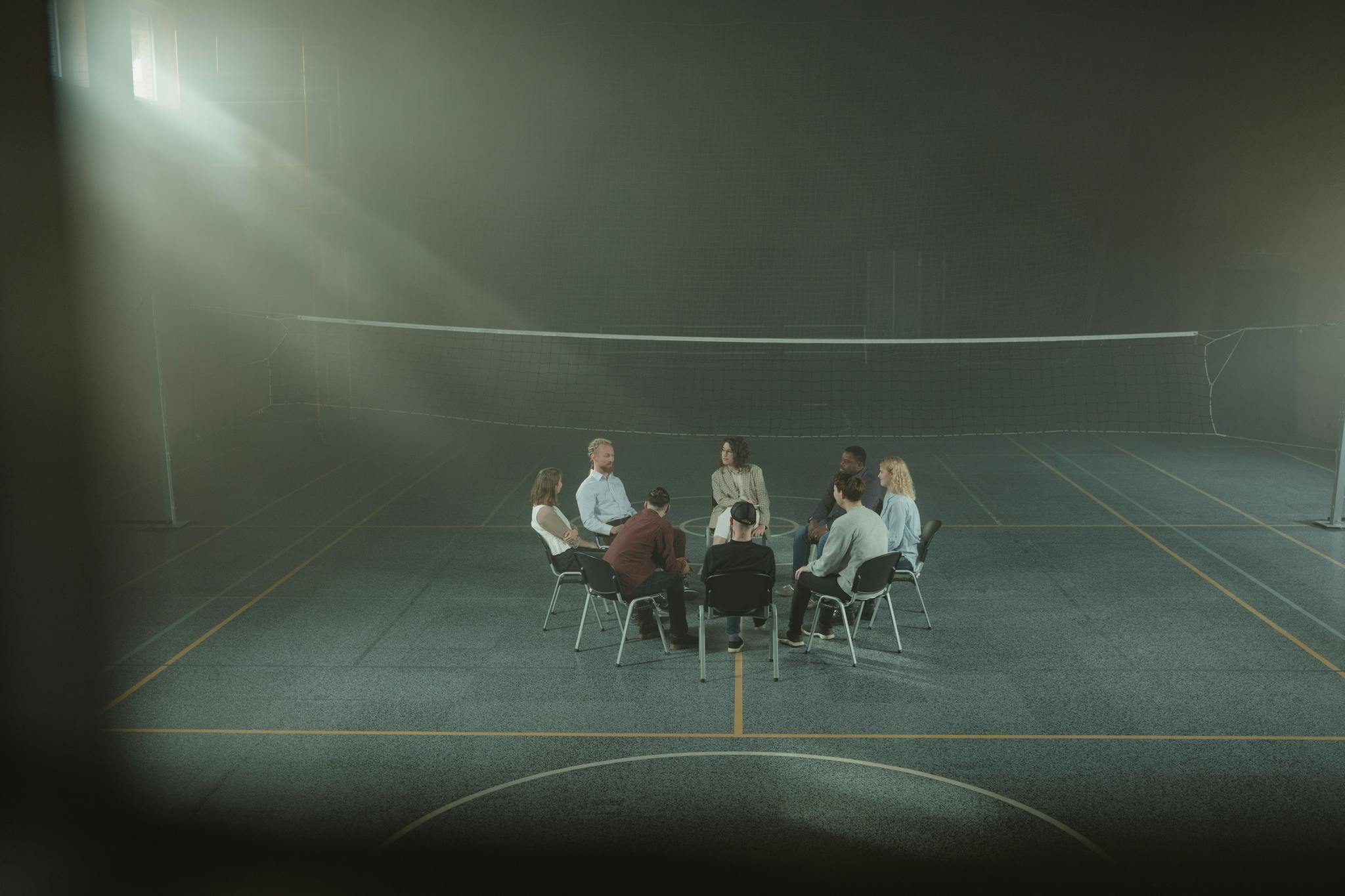 A group therapy session with participants seated in a circle inside a dimly lit gymnasium.